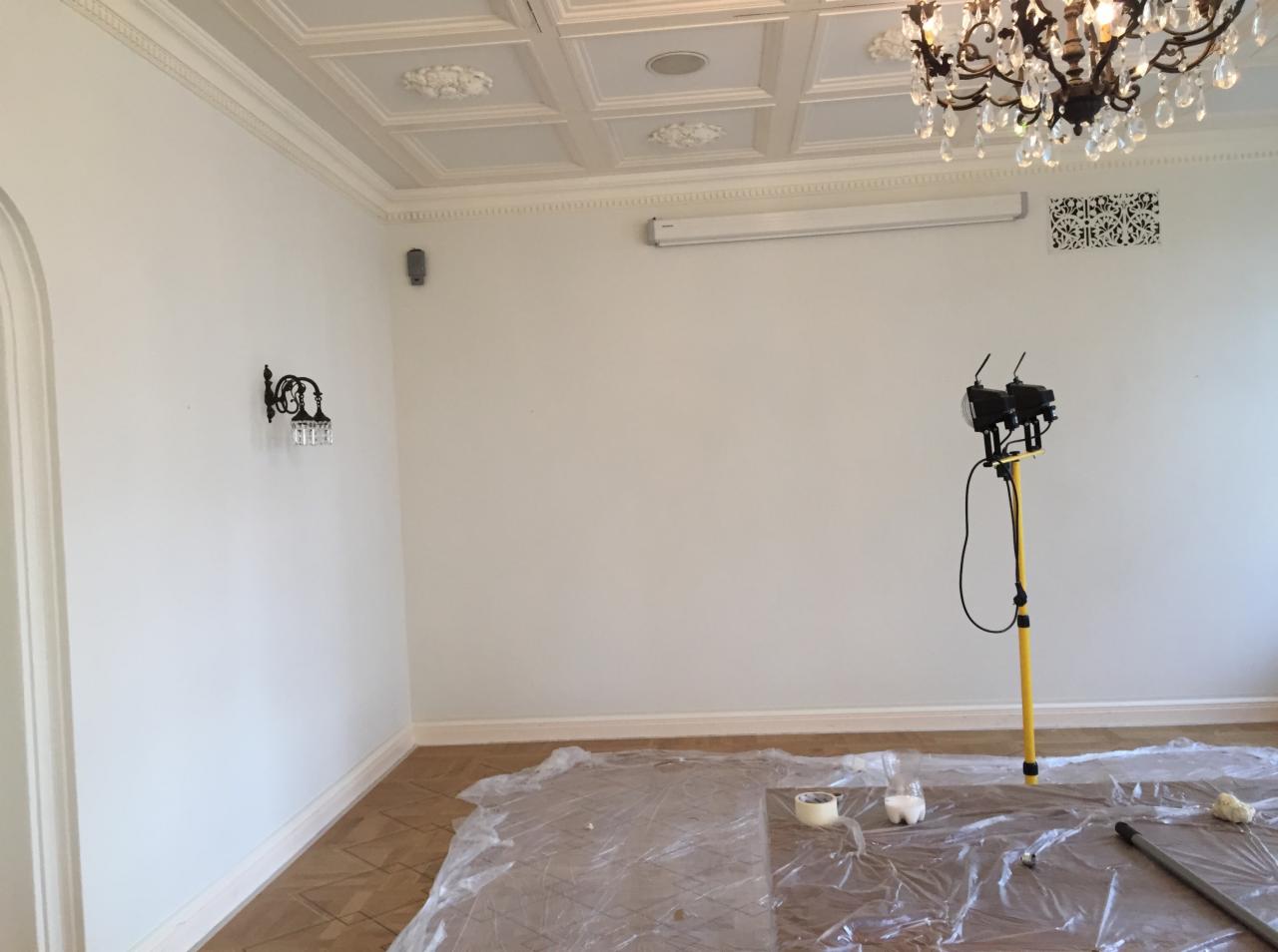 Pokój z dekoracyjnym sufitem i parkietem zabezpieczonym folią malarską, oświetlony lampą roboczą, przygotowany do prac wykończeniowych.