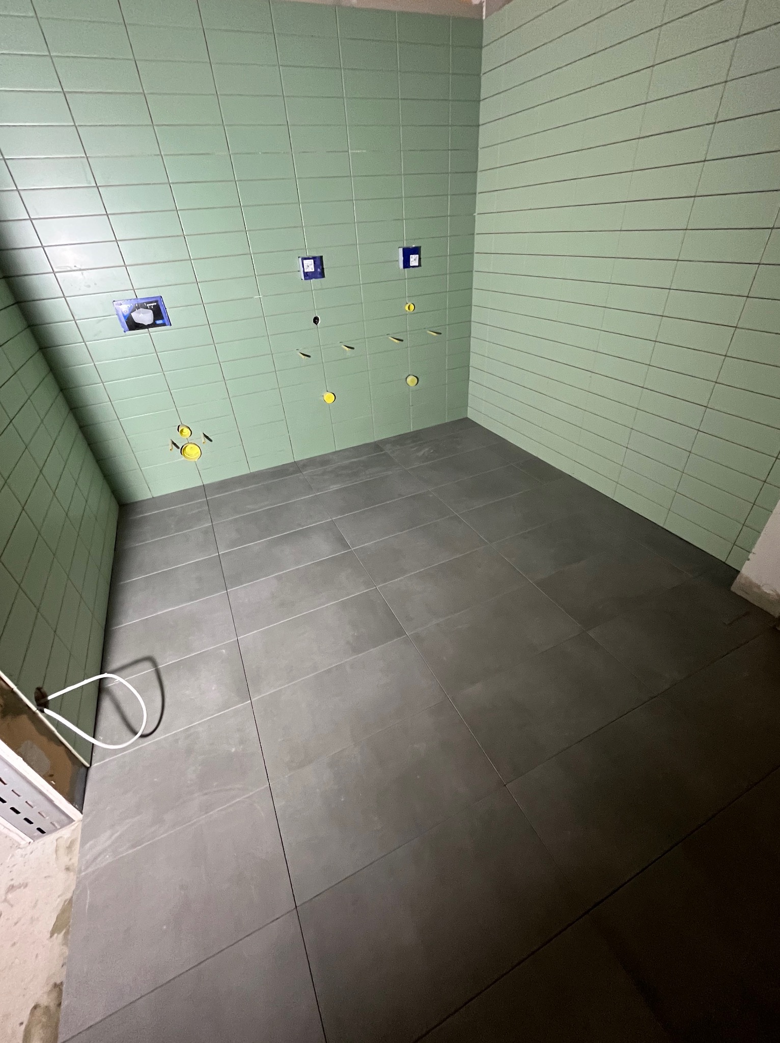 Świeżo wykafelkowana łazienka z jasnozielonymi, poziomymi płytkami ściennymi i dużymi, szarymi płytkami podłogowymi, widoczne otwory na instalacje wodne i kanalizacyjne.