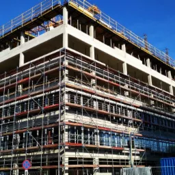 Wysoki budynek w trakcie budowy, otoczony rusztowaniami, z widocznym żurawiem w tle i niebieskim kontenerem na pierwszym planie, sfotografowany w słoneczny dzień.