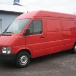 Czerwony samoch&oacute;d dostawczy zaparkowany na brukowanym placu, widoczny z boku, z budynkiem w tle.