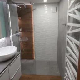 Nowoczesna łazienka po remoncie: biała umywalka nablatowa, kabina prysznicowa z drewnopodobnymi panelami i deszczownicą, ściany wykończone szarą i białą falistą płytką, grzejnik drabinkowy.