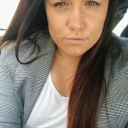 Portret kobiety z długimi, ciemnymi włosami i zielonymi oczami, ubrana w białą koszulkę i szarą marynarkę w kratę.