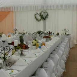 Uroczysta dekoracja stołu weselnego w białej tonacji z owocami, kwiatami i świecznikami, serwetki ułożone w trójkąty, krzesła z białymi pokrowcami, w tle dekoracja w kształcie dwóch serc z zieleni.