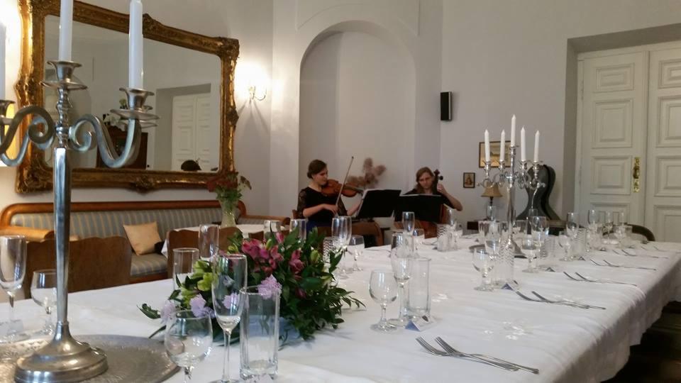 Uroczysty stół z białym obrusem, zastawą i kwiatami, w tle grający duet smyczkowy - skrzypaczka i wiolonczelistka, w przestronnym wnętrzu z lustrem w złotej ramie i białymi drzwiami.