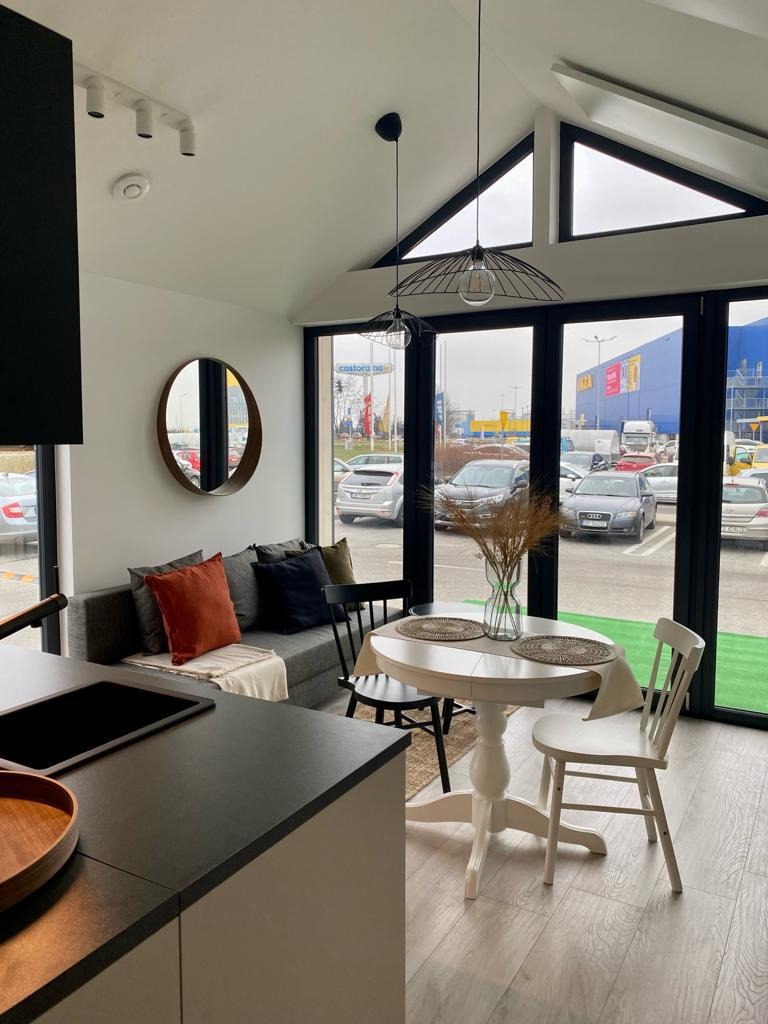 Wnętrze domu modułowego: jasna kuchnia z jadalnią, widok na parking zza okien. Minimalistyczny design, okrągły stół i czarne ramy okienne.