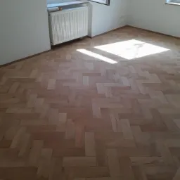 Parkiet ułożony w jodełkę w jasnym, pustym pokoju z białymi ścianami, oświetlony wpadającym przez okno światłem słonecznym.