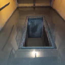 Świeżo wylana, mokra, szara posadzka z widocznym otworem inspekcyjnym w podłodze garażu lub warsztatu. Ściany żółte, w tle warsztatowy stół z narzędziami.