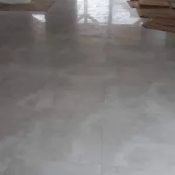 Jasnoszara, błyszcząca podłoga z płytek w trakcie remontu; widoczne kartony ochronne i taczka za szklanymi drzwiami.