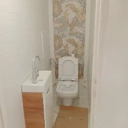 Mała łazienka z umywalką nablatową na szafce z drewnianym frontem i toaletą w tle, wykończona białymi płytkami z delikatną fakturą oraz dekoracyjnym pasem płytek z motywem liści w odcieniach...