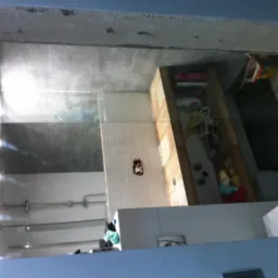 Wnętrze łazienki w trakcie prac wykończeniowych, widoczne białe płytki na ścianach, surowy betonowy sufit, instalacje sanitarne i elektryczne w trakcie montażu, oraz częściowo umeblowana przestrzeń...