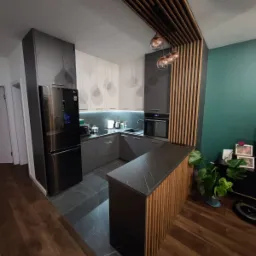 Nowoczesna kuchnia z szarymi i białymi szafkami, czarnym blatem, drewnianą zabudową sufitu i wyspą z drewnianymi lamelami, oświetleniem LED i wiszącymi lampami.