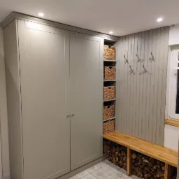 Szara szafa wnękowa z półkami na kosze wiklinowe, drewniana ławka z ułożonym pod spodem drewnem na opał, oraz wieszaki na ścianie w przedpokoju z wzorzystą podłogą.