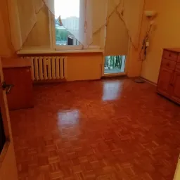 Pusta przestrzeń w mieszkaniu z parkietem w jodełkę, widok na okno z firankami i balkon, w tle budynki mieszkalne.