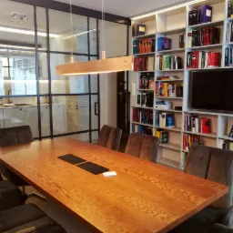 Sala konferencyjna z dużym drewnianym stołem, krzesłami z aksamitną tapicerką i regałem z książkami w tle, oddzielona szklaną ścianą od kuchni.