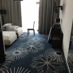 Pokój hotelowy w Łodzi z wzorzystą, niebieską wykładziną w motywy rozet, łóżkami i ciemnymi meblami.