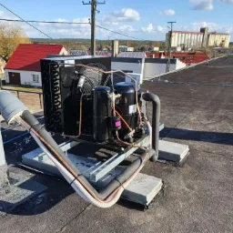 Kompaktowy agregat chłodniczy na dachu budynku, widoczne połączenia rurowe i elementy instalacji, w tle zabudowa miejska i błękitne niebo.