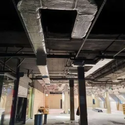 Widok z dołu na fragment systemu wentylacyjnego z izolacją termiczną, zamontowanego pod sufitem w dużym, pustym pomieszczeniu handlowym w trakcie remontu.