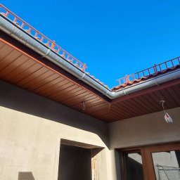 ArtHouse Artur Grzyb - Widok z dołu na fragment dachu z brązową podbitką, szarą rynną i czerwonymi dachówkami, na tle błękitnego nieba, ściana budynku w stanie surowym.