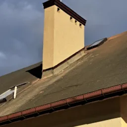 Beżowy komin z brązowym wykończeniem na dachu pokrytym papą, widoczne okno dachowe i rynna, na tle pochmurnego nieba.