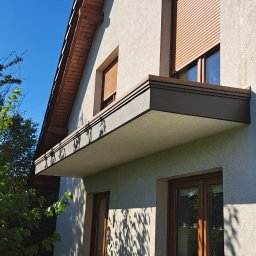 ArtHouse Artur Grzyb - Elewacja budynku z nowocześnie zabudowanym balkonem z brązowymi detalami, widok z dołu na tle błękitnego nieba i zieleni drzew.