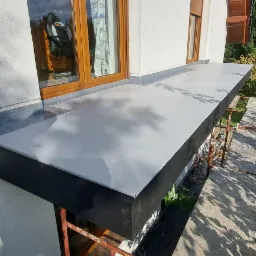 Wykończony balkon z szarą, gładką powierzchnią, widok z góry, rusztowanie w tle, okno z drewnianą ramą i zasłoną z motywem samolotów