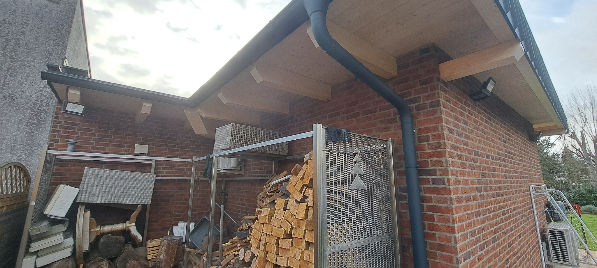 Zadaszenie nad składem drewna, widoczna konstrukcja drewniana, orynnowanie i murowana ściana budynku z oświetleniem zewnętrznym.