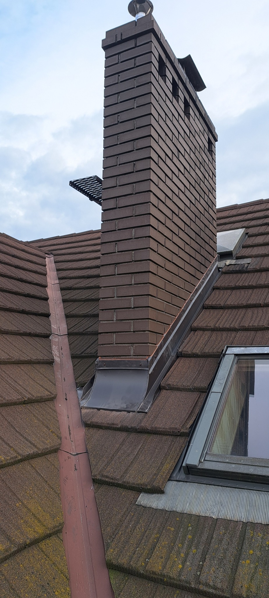 Widok z góry na brązowy dach z dachówki ceramicznej z kominem z cegły klinkierowej, obróbkami blacharskimi i oknem dachowym w pochmurny dzień.