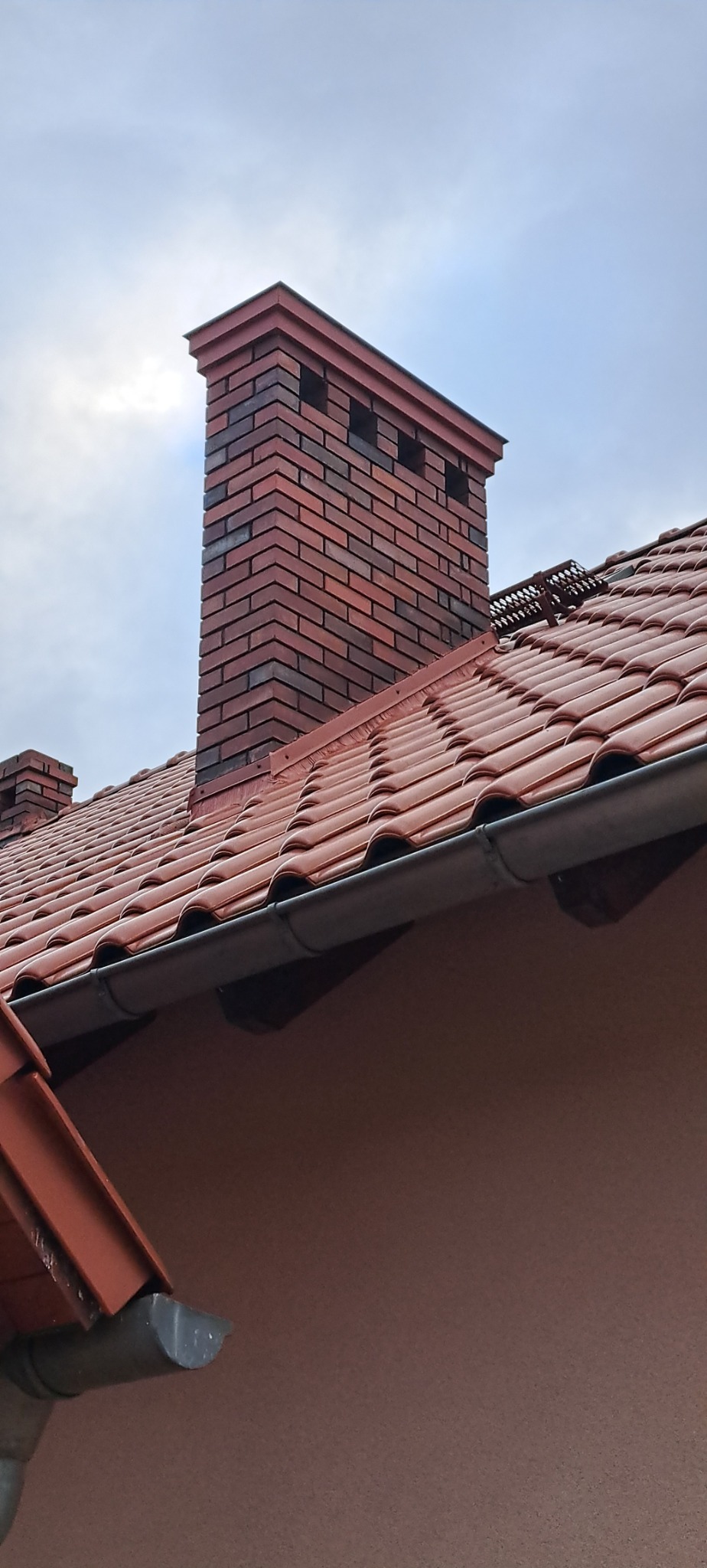 Nowy, ceglany komin z otworami wentylacyjnymi, wystający ponad czerwoną dachówkę na tle pochmurnego nieba. Widoczne również rynny i fragment elewacji budynku.