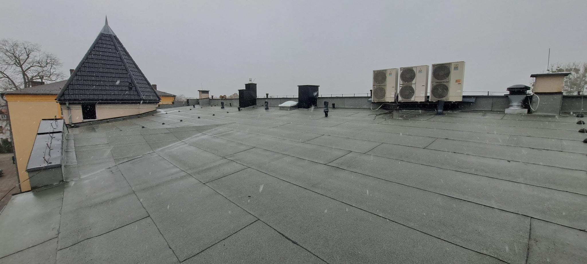 Szary, płaski dach pokryty papą bitumiczną w arkuszach, z widocznymi kominami, klimatyzatorami i wieżyczką z czarną dachówką w tle, podczas opadów śniegu.