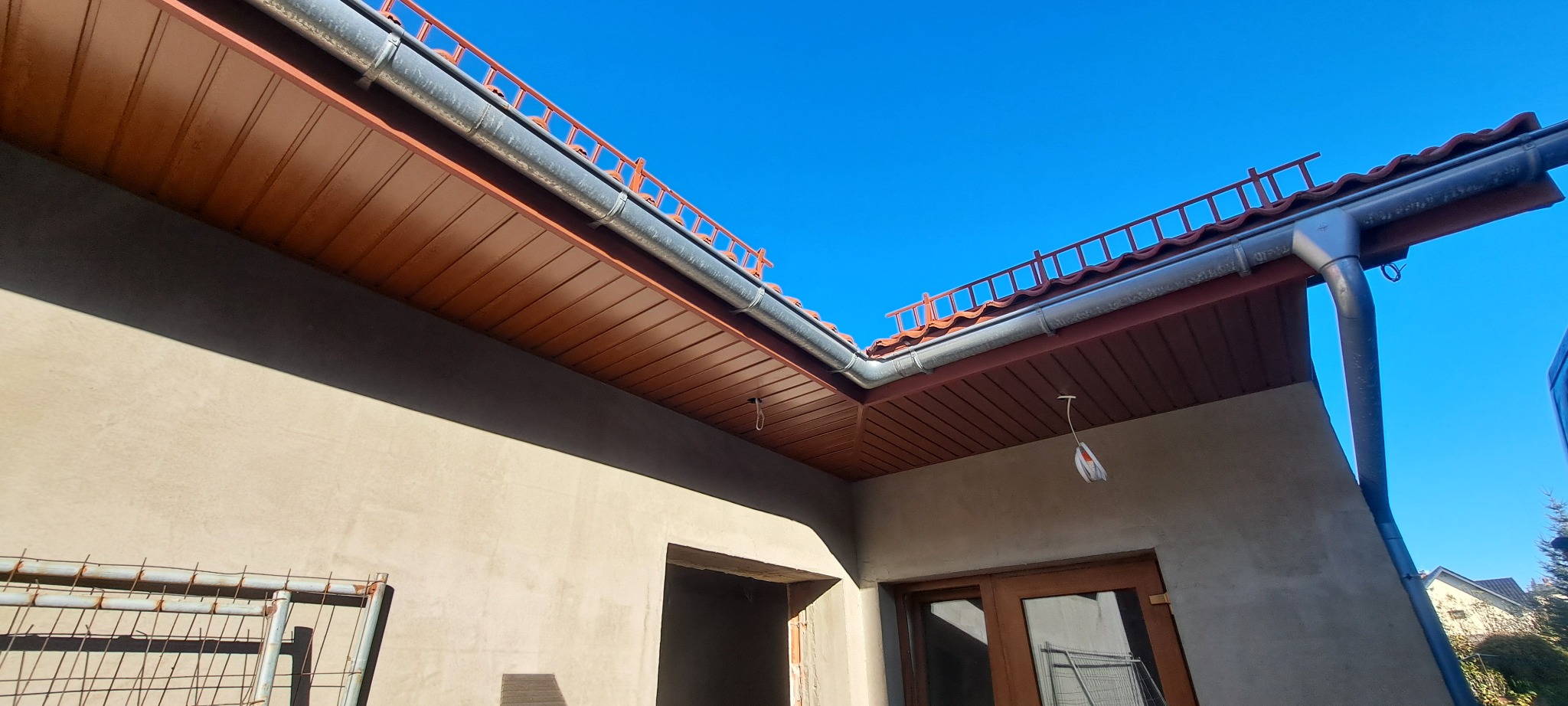 Widok z dołu na fragment dachu z brązową podbitką, szarą rynną i czerwonymi dachówkami, na tle błękitnego nieba, ściana budynku w stanie surowym.