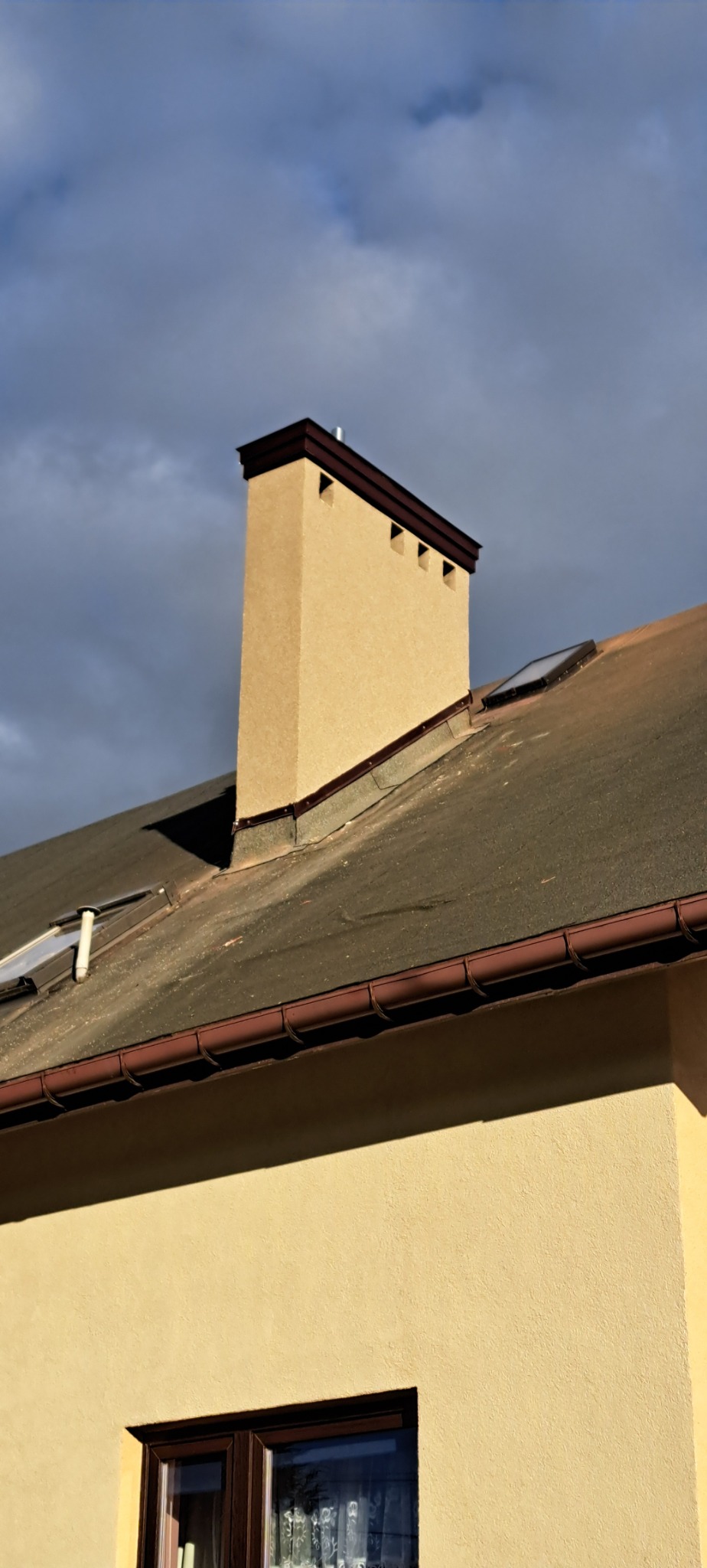 Beżowy komin z brązowym wykończeniem na dachu pokrytym papą, widoczne okno dachowe i rynna, na tle pochmurnego nieba.