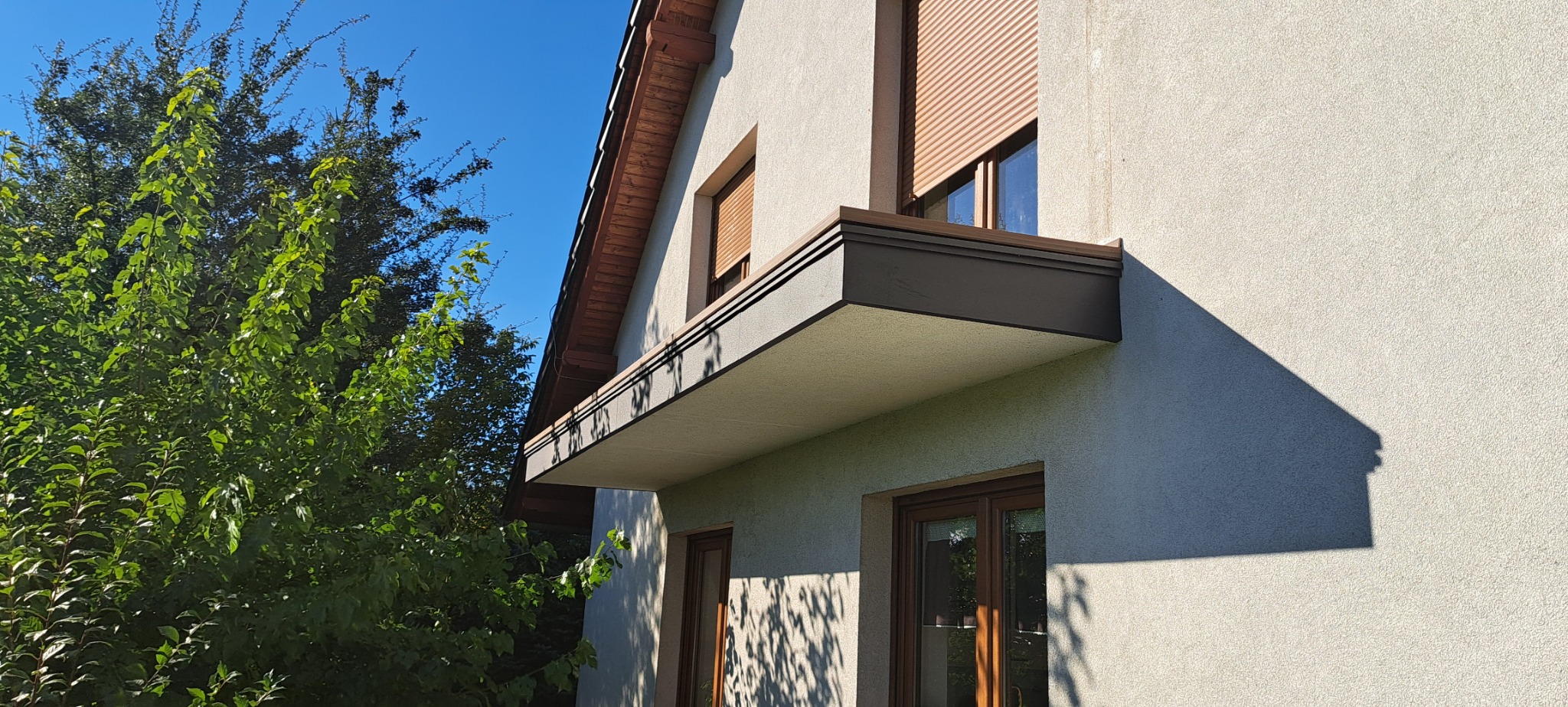 Elewacja budynku z nowocześnie zabudowanym balkonem z brązowymi detalami, widok z dołu na tle błękitnego nieba i zieleni drzew.