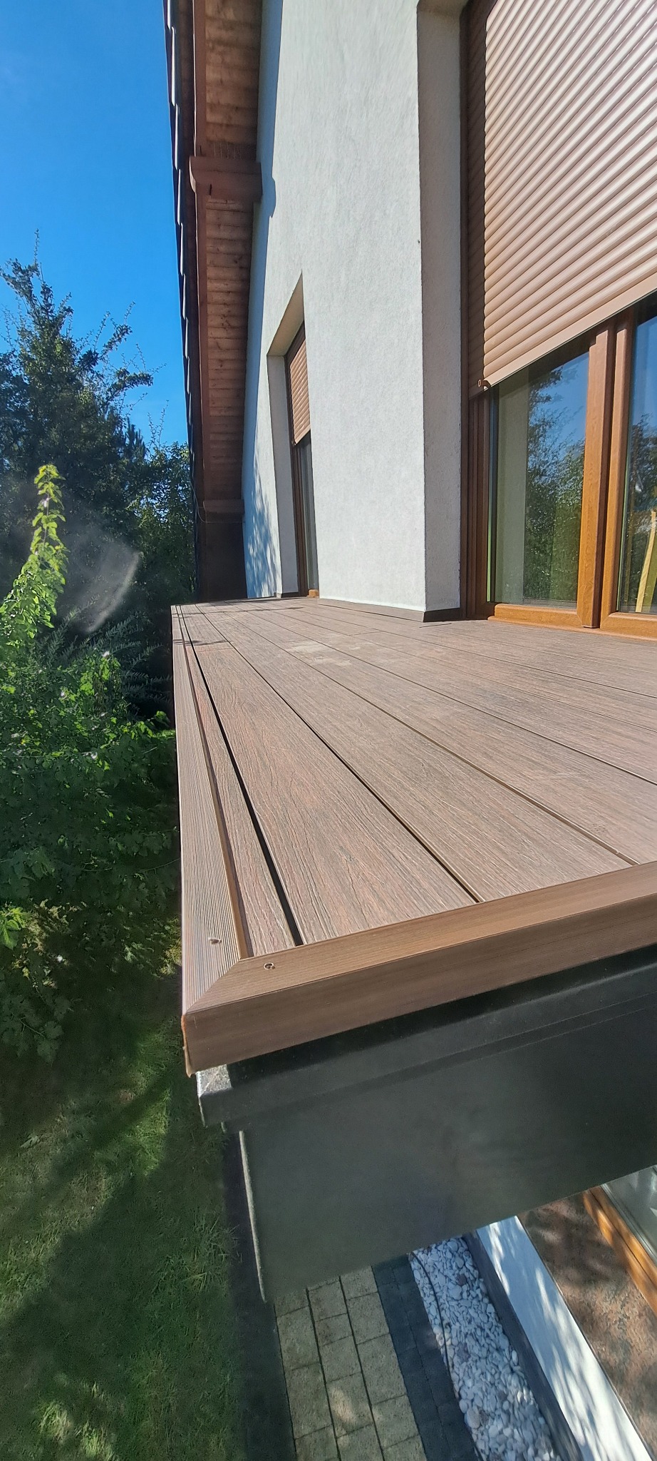 Nowoczesny balkon z kompozytowych desek w Lublińcu, widok z dołu, z widoczną elewacją budynku i roletami zewnętrznymi.