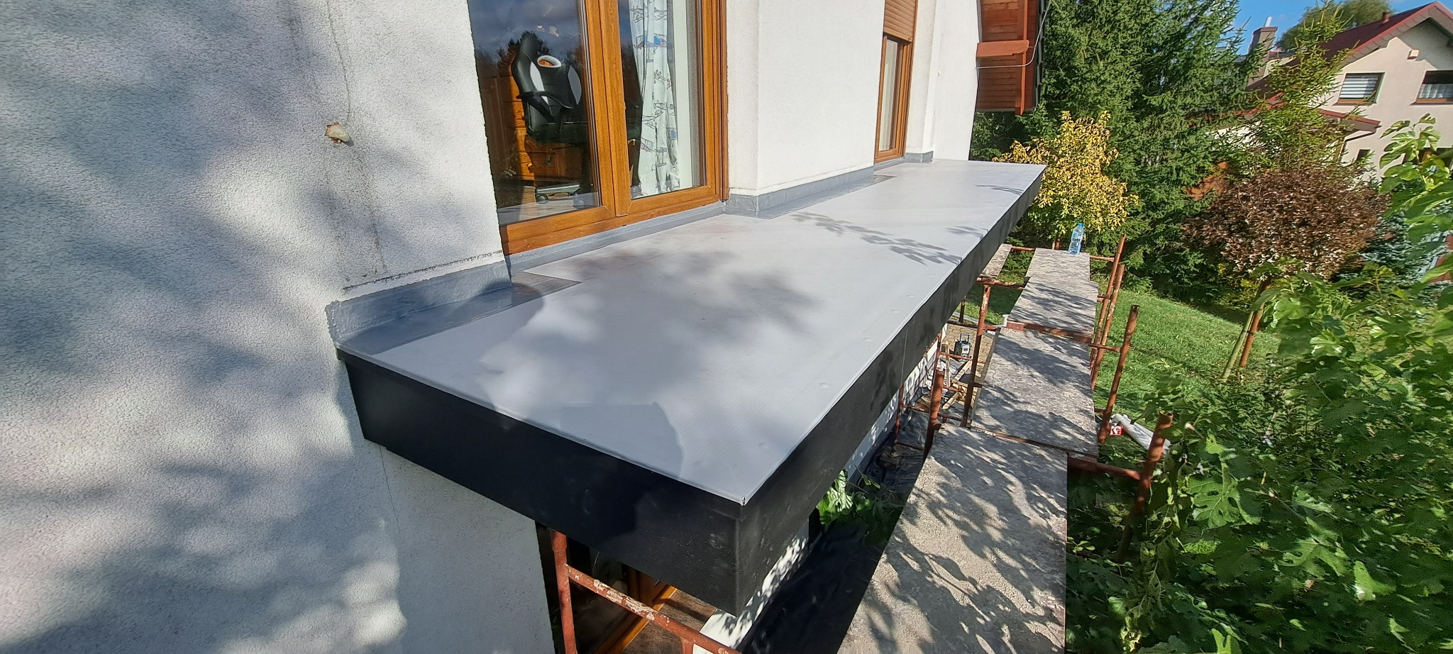 Wykończony balkon z szarą, gładką powierzchnią, widok z góry, rusztowanie w tle, okno z drewnianą ramą i zasłoną z motywem samolotów