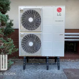 Zewnętrzna jednostka pompy ciepła LG ThermaV 12 kW na czarnym stelażu, zainstalowana przy ścianie budynku z ceglanym cokołem, obok zielonego krzewu.