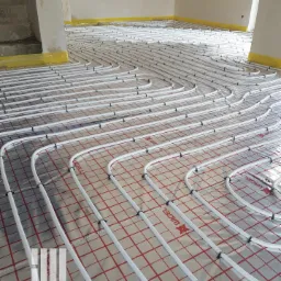 Białe rury ogrzewania podłogowego, mocowane czarnymi uchwytami, rozłożone na folii izolacyjnej z czerwonym nadrukiem siatki, w surowym wnętrzu z fragmentem schodów w tle.