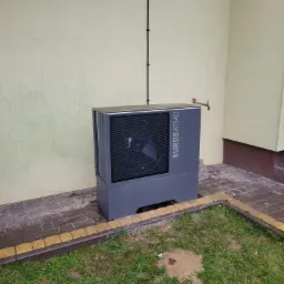 Szara pompa ciepła marki Eurosatmo zamontowana przy jasnej ścianie budynku, widoczna instalacja elektryczna i kran z wodą na zewnątrz.