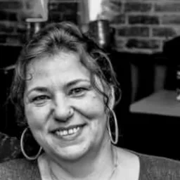 Portret kobiety z kolczykami-kołami i wisiorkiem w kształcie serca, na tle ceglanej ściany i dekoracji. Zdjęcie czarno-białe, z bliska.