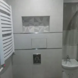 Wykończona łazienka w odcieniach szarości z grzejnikiem drabinkowym, umywalką z szafką i wnęką dekoracyjną z geometrycznym wzorem na ścianie.