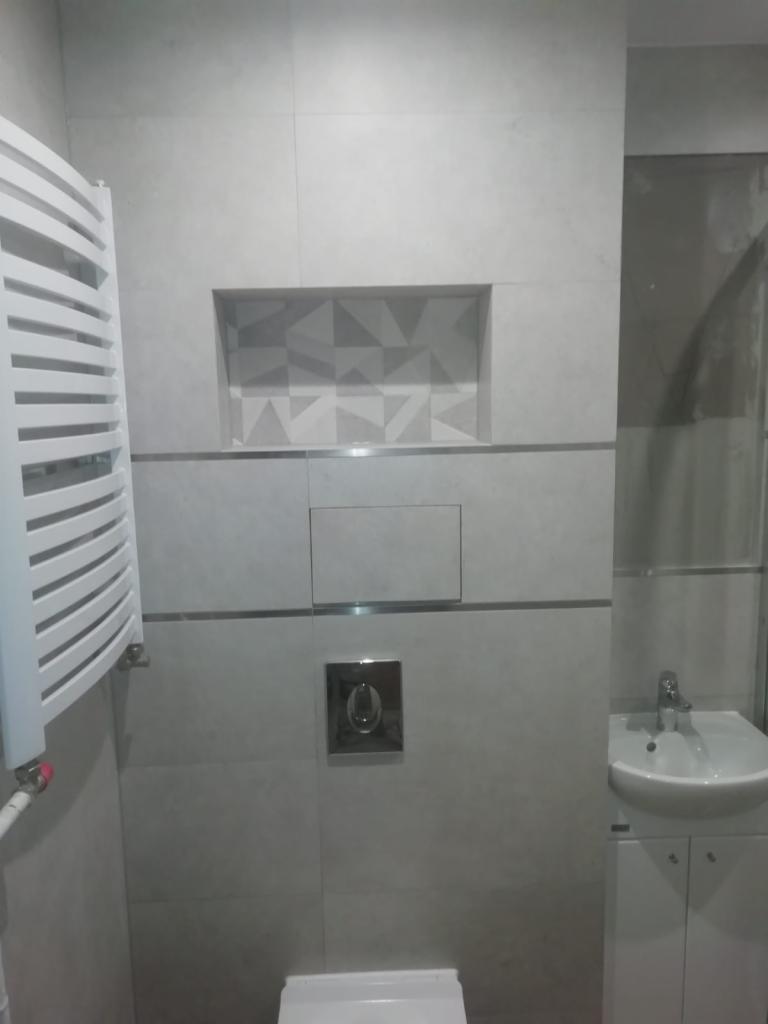 Wykończona łazienka w odcieniach szarości z grzejnikiem drabinkowym, umywalką z szafką i wnęką dekoracyjną z geometrycznym wzorem na ścianie.