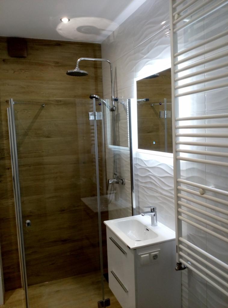 Nowoczesna łazienka z kabiną prysznicową ze szklanymi drzwiami, umywalką nablatową na szafce, podświetlanym lustrem i grzejnikiem drabinkowym; ściany wykończone płytkami imitującymi drewno...