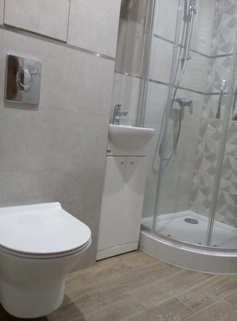 Nowoczesna łazienka z kabiną prysznicową, umywalką z szafką i podwieszaną toaletą, wykończona szarymi płytkami z metalowymi listwami i podłogą imitującą drewno.