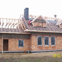 Budowa domu jednorodzinnego w trakcie: widoczna murowana konstrukcja ścian, drewniana więźba dachowa w trakcie montażu, komin oraz pracownik w czerwonym kombinezonie na dachu.