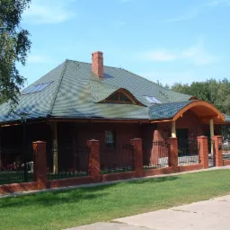 Dom jednorodzinny z zielonym dachem z dachówką karpiówką, ceglanym kominem i oknami dachowymi, otoczony ceglanym ogrodzeniem z kutymi elementami.