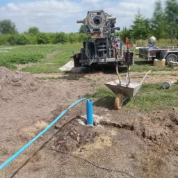 Wiertnica do studni głębinowych na działce, widoczny niebieski rurociąg, taczka z drobnym żwirem i przyczepka w tle.