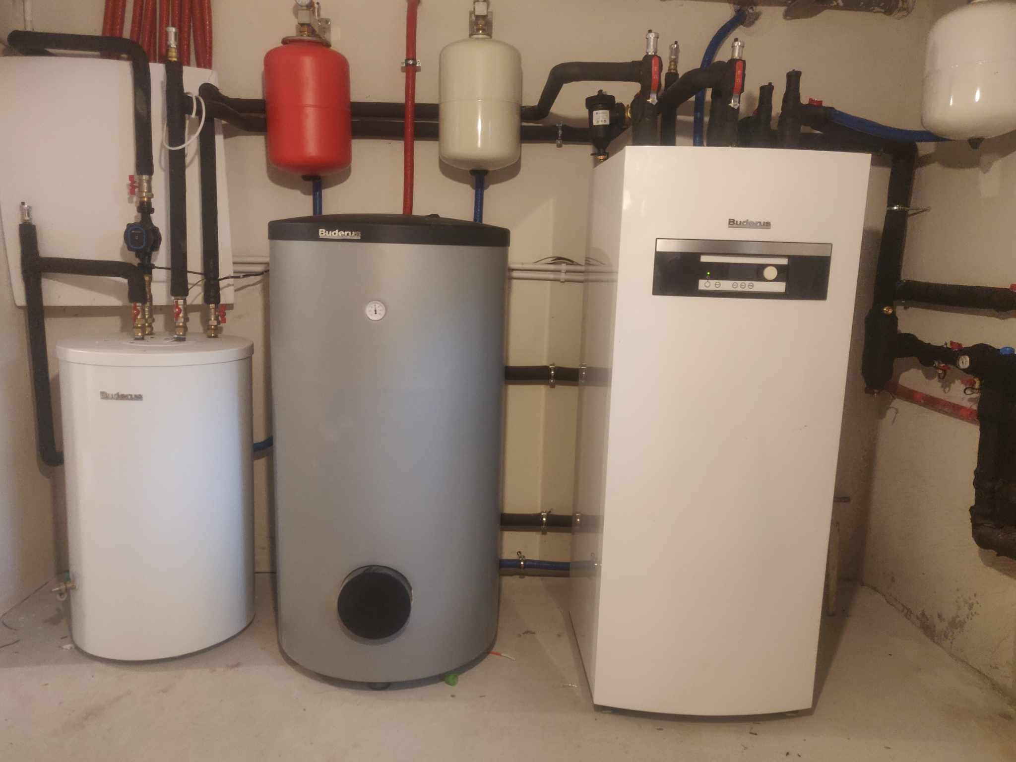 Kotłownia z zainstalowanym piecem gazowym Buderus, dwoma zbiornikami buforowymi (czerwonym i szarym) oraz zasobnikiem ciepłej wody użytkowej, widoczne rury instalacji CO i CWU.