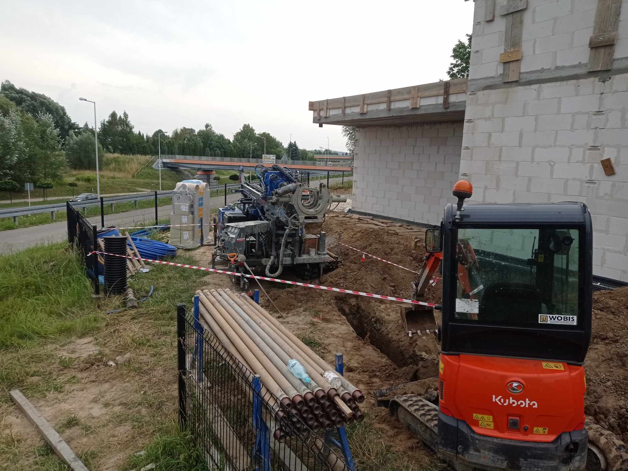 Wiertnica do studni głębinowych na placu budowy, obok wykopu i mini-koparki Kubota, rury wiertnicze ułożone na ogrodzeniu, w tle widoczny most.