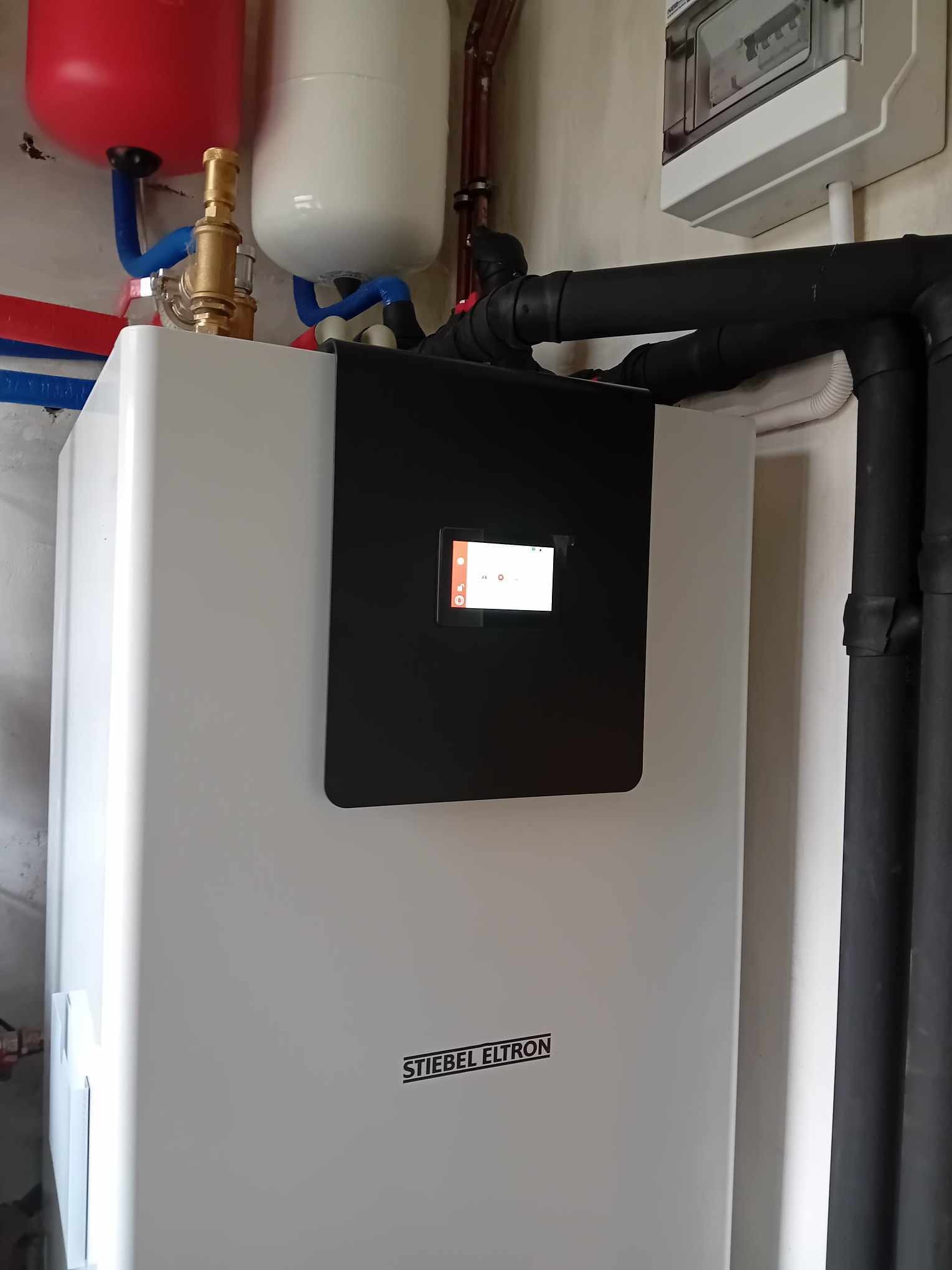 Biała pompa ciepła marki Stiebel Eltron z czarnym panelem sterowania, podłączona do instalacji z rurami w izolacji, widoczne zbiorniki wyrównawcze i skrzynka z bezpiecznikami.