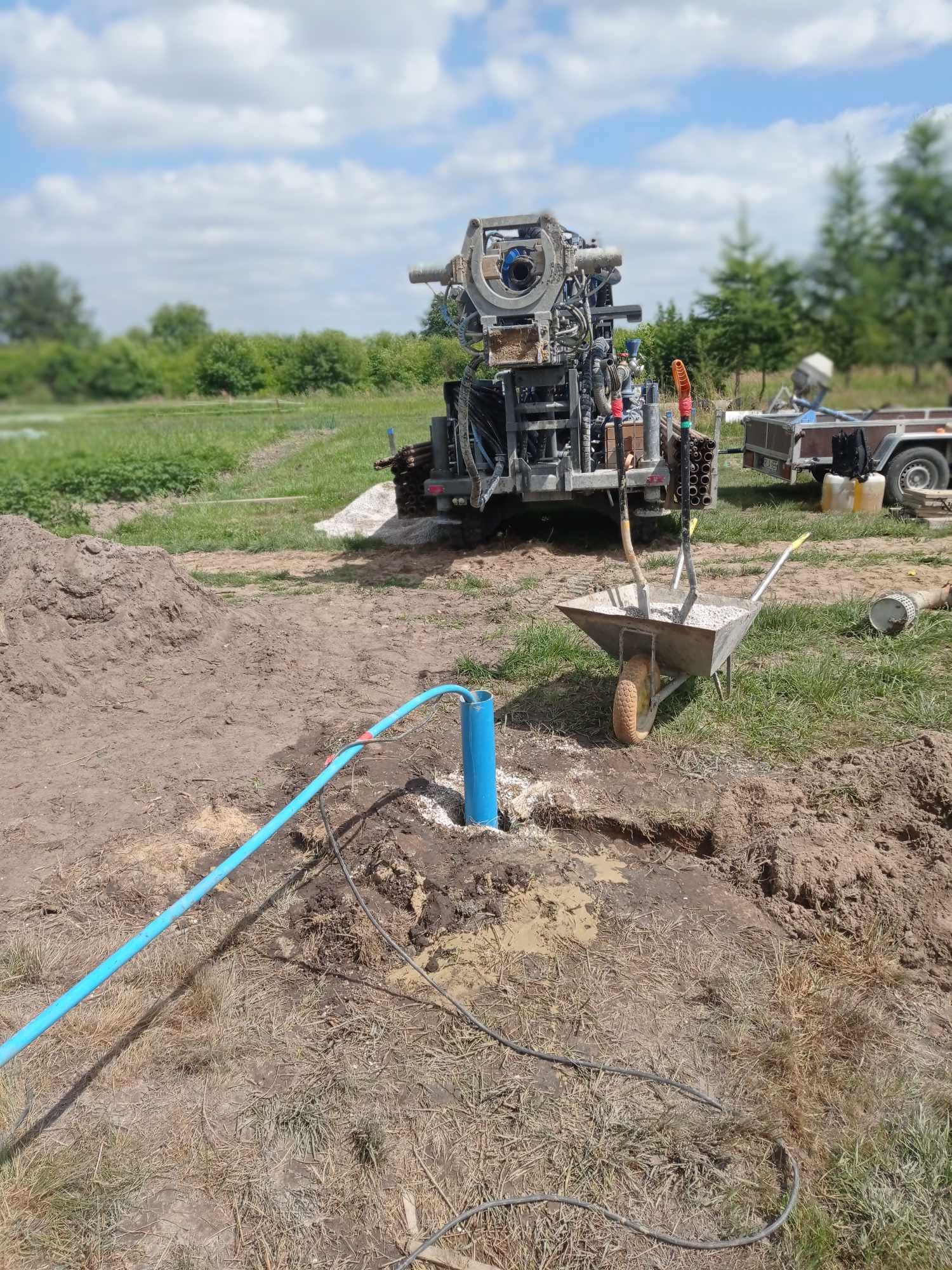 Wiertnica do studni głębinowych na działce, widoczny niebieski rurociąg, taczka z drobnym żwirem i przyczepka w tle.