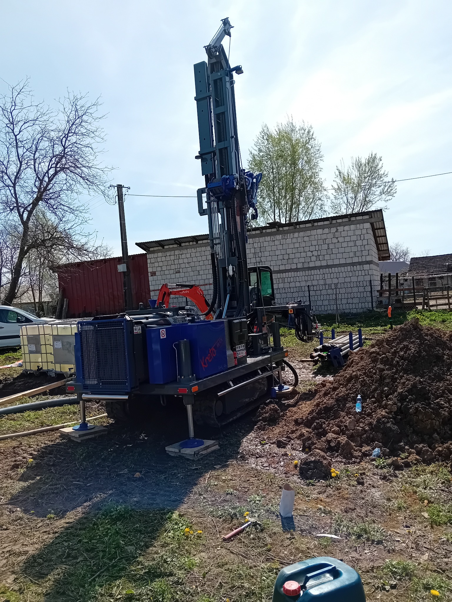 Wiertnica do studni głębinowych marki Kreto VHL460 na gąsienicach, z widocznym wykopem, zbiornikami na wodę i stertą ziemi, w tle budynek gospodarczy z białej cegły i fragment krajobrazu wiejskiego.